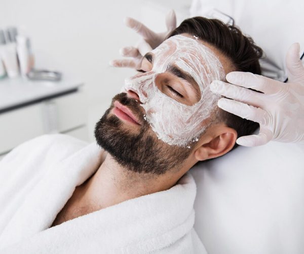 Calm bearded man lying with closed eyes and having white clay mask on the face while cosmetologist touching his temples
